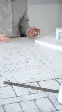Mimarlar detaylı planlar ve beyaz bir masanın üzerinde bir ölçek modeli etrafında toplanarak, modern bir konut projesinde işbirliği yaparken planları analiz ediyor, araçlar ve belgeler çiziyor ve kapatıyorlar.