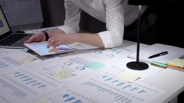 Meşgul profesyonel yönetici, çok sayıda finansal raporu ve iş belgelerini aktif olarak analiz ederek, bir dizüstü bilgisayar ve kalemle istatistikleri gözden geçirmek için, parlak bir şekilde aydınlatılmış bir masa üzerinde çalışmaktadır.