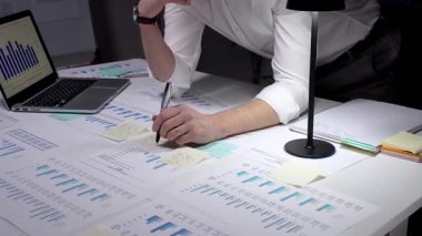 İş adamı finansal verileri ve istatistikleri analiz ediyor, ekranda grafikler ve çizelgeler olan bir dizüstü bilgisayarda yazıyor ve loş bir ofiste masaya yayılmış raporları inceliyor.