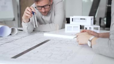 Bir konut projesinde işbirliği yapan mimarlar, planlar ve ölçekli bir modeli bir masada inceleyerek, tasarım detaylarını, inşaat planlarını ve gelecekteki gelişimi tartışarak,