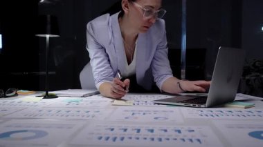 Finansal raporlar ve çizelgelerle kaplı bir masada duran bir kadın masa lambasının yumuşak parıltısı altında özenle bir dizüstü bilgisayar üzerinde çalışıyor, kendini işine adamış ve iş verilerinin detaylı analizini yapıyor.