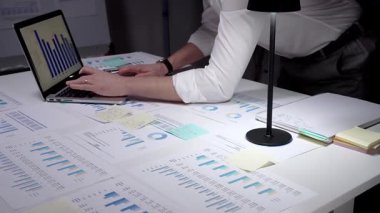İş adamı finansal verileri ve istatistikleri analiz ediyor, ekranda grafikler ve çizelgeler olan bir dizüstü bilgisayarda yazıyor ve loş bir ofiste masaya yayılmış raporları inceliyor.
