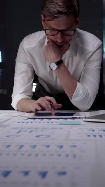 Profesyonel iş adamı masaya yaslanıyor. Gece geç saatlerde finansal veri ve çizelgeleri analiz ediyorum. Loş bir ofis ortamında fazla mesai yapmak. Ayrıntılı raporlara ve stratejik planlamaya odaklanmış