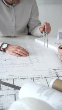 Mimarlar detaylı planlar ve beyaz bir masanın üzerinde bir ölçek modeli etrafında toplanarak, modern bir konut projesinde işbirliği yaparken planları analiz ediyor, araçlar ve belgeler çiziyor ve kapatıyorlar.