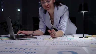 Finansal raporlar ve çizelgelerle kaplı bir masada duran bir kadın masa lambasının yumuşak parıltısı altında özenle bir dizüstü bilgisayar üzerinde çalışıyor, kendini işine adamış ve iş verilerinin detaylı analizini yapıyor.