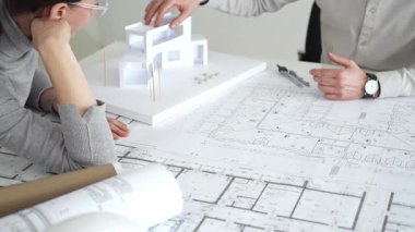 İki mimar yeni bir bina tasarımında işbirliği yapıyor ayrıntılı planları gözden geçiriyorlar ve pusula ile beyaz bir mimari modelin yanındaki planları titizlikle ölçüyorlar.