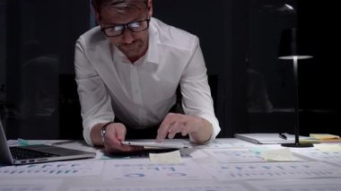 Gece geç saatlere kadar karanlık bir ofiste çalışan profesyonel işadamı, mali verilere odaklanıyor, bir tablet, çizelgeler ve raporlar kullanarak teslim tarihleri, fazla mesaileri ve çalışkan çalışma ahlakını yansıtıyor