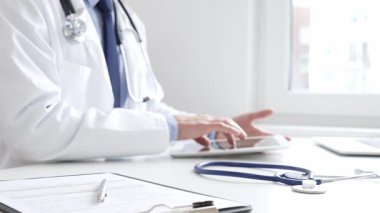 Klinikte dijital tablet kullanan doktor adamın yanındaki masada mavi steteskop duruyor. Tıp ve sağlık hizmetleri kavramı