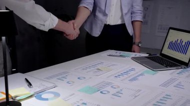 Profesyonel giysiler içinde görünmeyen iki iş adamı el sıkışıyor, mali tabloları ve verileri gece modern ofis ortamında gözden geçirdikten sonra önemli bir anlaşmaya varıyorlar.