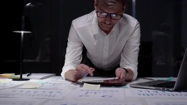 Gece geç saatlere kadar karanlık bir ofiste çalışan profesyonel işadamı, mali verilere odaklanıyor, bir tablet, çizelgeler ve raporlar kullanarak teslim tarihleri, fazla mesaileri ve çalışkan çalışma ahlakını yansıtıyor