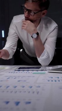 Profesyonel iş adamı masaya yaslanıyor. Gece geç saatlerde finansal veri ve çizelgeleri analiz ediyorum. Loş bir ofis ortamında fazla mesai yapmak. Ayrıntılı raporlara ve stratejik planlamaya odaklanmış