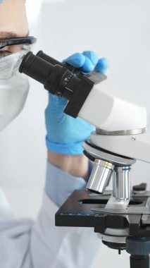 Mavi koruyucu giysiler giyen kadın bilim adamı mikroskop kullanarak laboratuvar ortamında örnekler inceleyerek bilimsel araştırma ve analiz yapıyor. Tıp ve bilim konsepti.