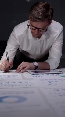 Profesyonel iş adamı masaya yaslanıyor. Gece geç saatlerde finansal veri ve çizelgeleri analiz ediyorum. Loş bir ofis ortamında fazla mesai yapmak. Ayrıntılı raporlara ve stratejik planlamaya odaklanmış