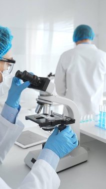 Laboratuvar önlüklü, yüz maskeli ve koruyucu gözlüklü iki bilim adamı laboratuarda mikroskop kullanarak araştırma yapıyorlar. Tıp ve bilim konsepti.