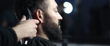 Moda bir berber dükkanında elektrikli makasla saç kesen profesyonel berber sakallı bir adam için modern bir saç stili yapıyor.