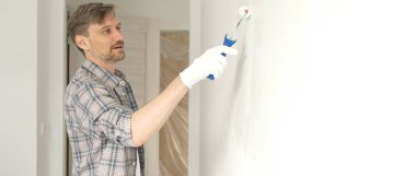 Kareli gömlek giyen profesyonel erkek ressam, apartman duvarına beyaz boya sürüyor. Kapsamlı ev tadilatı sırasında düzenli darbeler alarak iç mekanı değiştiriyor..