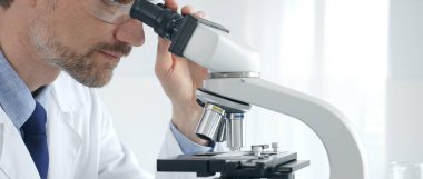 Yetişkin erkek bilim araştırmacısı mikroskopla örnek analiz ediyor, yüksek teknoloji laboratuvar ortamında koruyucu giysiler giyiyor. Sağlık, tıp ve bilim konsepti.