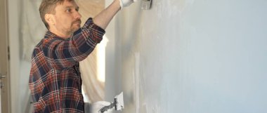 Eldiven giyen erkek inşaat işçisi, bir el aleti kullanarak duvara alçı yapıştırıyor, ev yenileme veya inşaat projesine katkıda bulunuyor