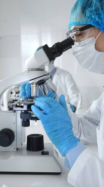 Bilinmeyen bilim adamı mavi güvenlik donanımı takıyor. Araştırma için mikroskop kullanıyor. Erkek bir meslektaşı arka planda çalışıyor. Tıp, sağlık ve bilim konsepti.