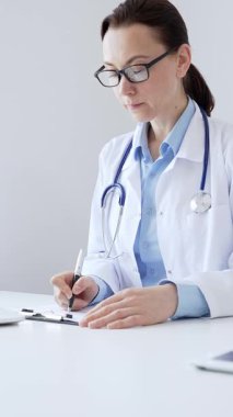 Orta yaşlı kadın doktor, sağlık evraklarını tamamlıyor, dizüstü bilgisayarın yanına yerleştirilmiş bir pano ve steteskop, pencerenin yanında iyi aydınlatılmış klinik çalışma alanı. İlaç ve sağlık hizmetleri.