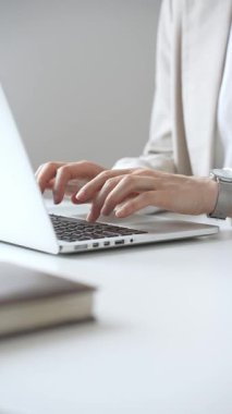 Şık bej bir takım elbise giymiş, gümüş bir saat takan, parlak beyaz bir masada oturan, dizüstü bilgisayara odaklanmış, işine odaklanmış bir iş kadını. İş adamları konsepti