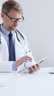 Laboratuvar önlüğü ve steteskop giyen orta yaşlı doktor, tıp bürosunda masasında otururken dijital tablet kullanıyor, ön planda da da tıbbi çizelge ve kalem var. Tıp konsepti.