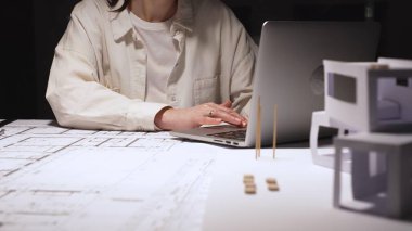 Genç profesyonel mimar veya mühendis, bir masada bina planları ve yapısal planlar üzerinde çalışır, teknik çizimler ve hesaplamaları incelerken dizüstü bilgisayarda yazar.