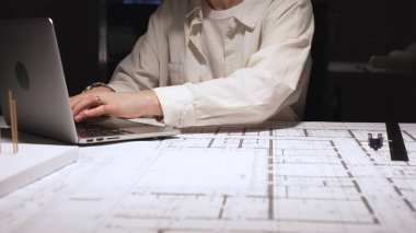 Genç profesyonel mimar veya mühendis, bir masada bina planları ve yapısal planlar üzerinde çalışır, teknik çizimler ve hesaplamaları incelerken dizüstü bilgisayarda yazar.