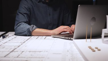 Dizüstü bilgisayar üzerinde çalışan tasarımcı ya da mimar, iyi aydınlatılmış bir masa üzerinde fiziksel modeller çiziyor, özveriyi, hassasiyeti ve yoğun bir profesyonel ortamı temsil ediyor.