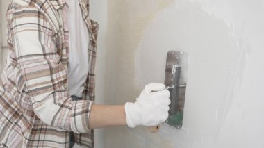 Eldiven giyen kimliği belirsiz kadın inşaat işçisi duvara sıva uygulamak için macun bıçağı kullanıyor. Ev yenileme projesi sırasında yüzeyi düzleştiriyor.