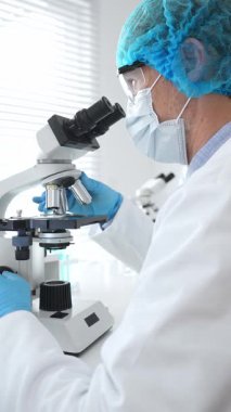 Koruyucu maske, şapka ve eldiven takan kıdemli erkek bilim adamı modern bir laboratuvarda mikroskop altında örnekleri inceliyor. Tıp, sağlık ve bilim konsepti.