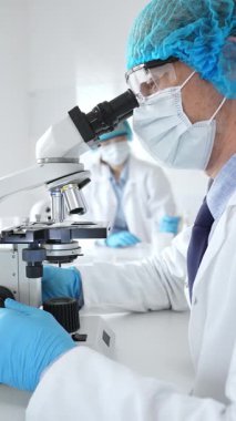 Koruyucu maske, şapka ve eldiven takan kıdemli erkek bilim adamı modern bir laboratuvarda mikroskop altında örnekleri inceliyor. Tıp, sağlık ve bilim konsepti.