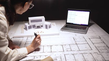 Female architect creating new construction project, drawing detailed architectural plans and designing innovative structures with a model and digital tools in a professional workspace