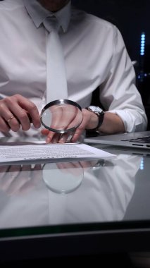 Bir belgeyi büyüteç kullanarak inceleyen, geç saatlere kadar çalışan, yasal bir anlaşmada ince baskı ve şartlara odaklanan bir iş adamı. Denetim ve aşırı çalışma kavramları.