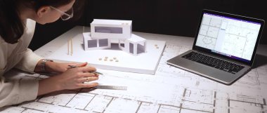 Mimar, mimari tasarım ve yapısal mühendislik üzerine cetvel ve kalemle, kaba yazılım ve fiziksel ölçekli bir bilgisayar kullanarak, bir binanın planına kesin çizgiler çiziyor.