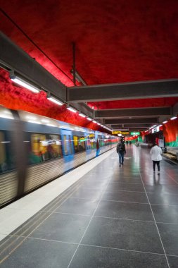 Stockholm, İsveç 'teki tren istasyonunda. Kırmızı ışıkla aydınlatılan kayaların altında platformda yürüyen birkaç yolcu var..
