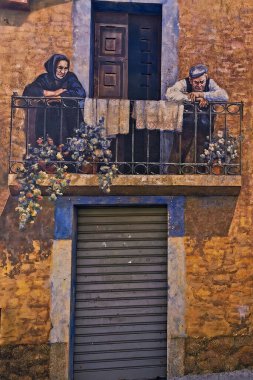 Balkonda yaşlı bir adam ve parmaklıklara yaslanmış bir kadın var. Bina cephesinde sanatsal boya. Fonni, Sardinya, İtalya 'dan ekran görüntüsü.