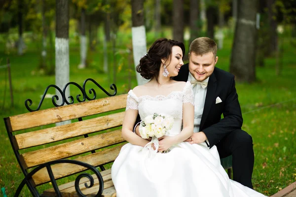 Día de la boda: hermosa novia y novio sentarse en el banco en el parque —  Fotos de Stock