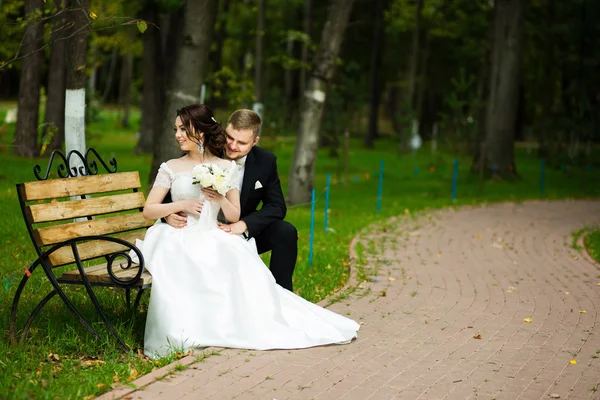 Esküvő napján: a gyönyörű menyasszony és a vőlegény ül a padon a parkban — Stock Fotó