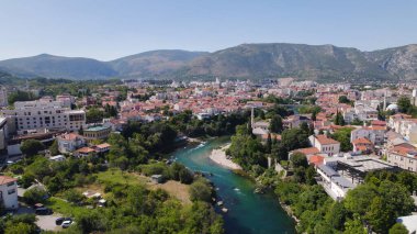 Mostar 'ın şehir manzarasını gösteren insansız hava aracı fotoğrafı, Neretva Nehri' nin tarihi eski kasaba ve çevre binalardan aktığını, manzaralı Balkan tepeleri tarafından çerçevelendiğini gösteriyor..