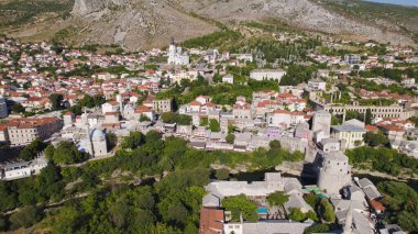 Mostar şehrinin arka planda yükselen Kutsal Üçlü Katedrali ile birlikte tarihi binalar, Neretva Nehri ve manzaralı Balkan tepeleriyle çevrili insansız hava aracı fotoğrafı..
