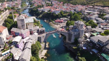 Mostar 'daki Neretva Nehri üzerinde tarihi binalarla birlikte Stari ikonik köprüsünün altından geçen küçük bir tekneyi gösteren insansız hava aracı fotoğrafı.
