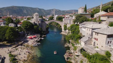 Mostar 'daki turkuaz Neretva Nehri boyunca tarihi binalarla çevrili Stari Köprüsü' nün altından geçen küçük bir tekneyi gösteren insansız hava aracı fotoğrafı..