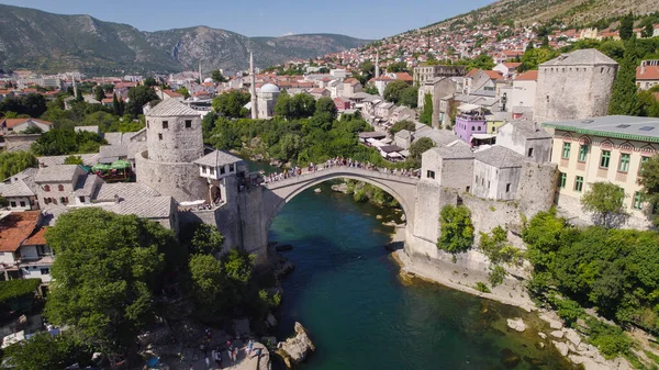 İnsansız hava aracı fotoğrafı, Stari Most köprüsünde, tarihi Mostar kenti ve arka planda renkli evler bulunan Neretva Nehri üzerindeki ziyaretçileri gösteriyor..