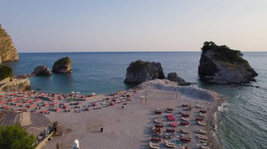 Karadağ 'ın Budva kentindeki Sveti Nikola Adası yakınlarındaki sakin bir plajın insansız hava aracı fotoğrafı, sakin turkuaz suların üzerinde sıcak alacakaranlık ışığında yıkandı..