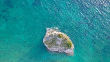 Havadan aşağıya doğru çekilen hava aracı fotoğrafı, göz kamaştırıcı turkuaz mavi sularla çevrili ıssız bir kayayı yakalıyor. Kristal berrak deniz sualtı dokularını ve gradyanlarını ortaya çıkararak canlı okyanus renkleri ve doğal güzelliği ile sakin ve minimal bir deniz manzarası yaratır.