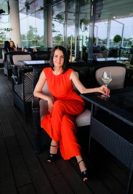 Güzel bir kadın şarap kadehiyle restoran terasında dinleniyor. Moda, esmer model, kırmızı takım elbise giyiyor..
