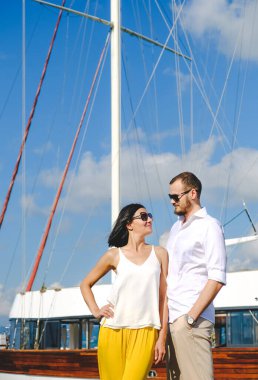 Limanda yürüyen mutlu aşıklar çifti. Arka planda gemi var. Beyaz bluzlu, neşeli, gülümseyen adam ve beyaz bluzlu şanslı kadın ve sarı pantolon, açık deniz limanında..