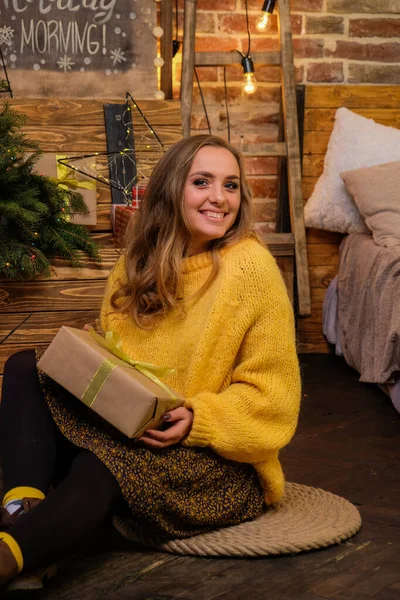 Portrait of happy, attractive woman with christmas gift on floor at home. Loft style christmas cozy decor on the background. Blonde, cute girl in fluffy, yellow oversized sweater. New Year\'s eve mood.