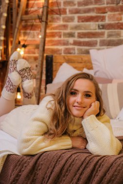 Evdeki yatakta gülümseyen genç bir kadın. Arka planda çatı katı tarzı Noel dekorasyonu var. Mutlu Noeller ve mutlu tatiller. Sarışın, tatlı, beyaz kazaklı ve İskandinav çoraplı bir kadın. Yeni yıl arifesi havası..